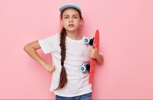 Stylish young woman in a light blue cap holding a skateboard against a solid pink background.