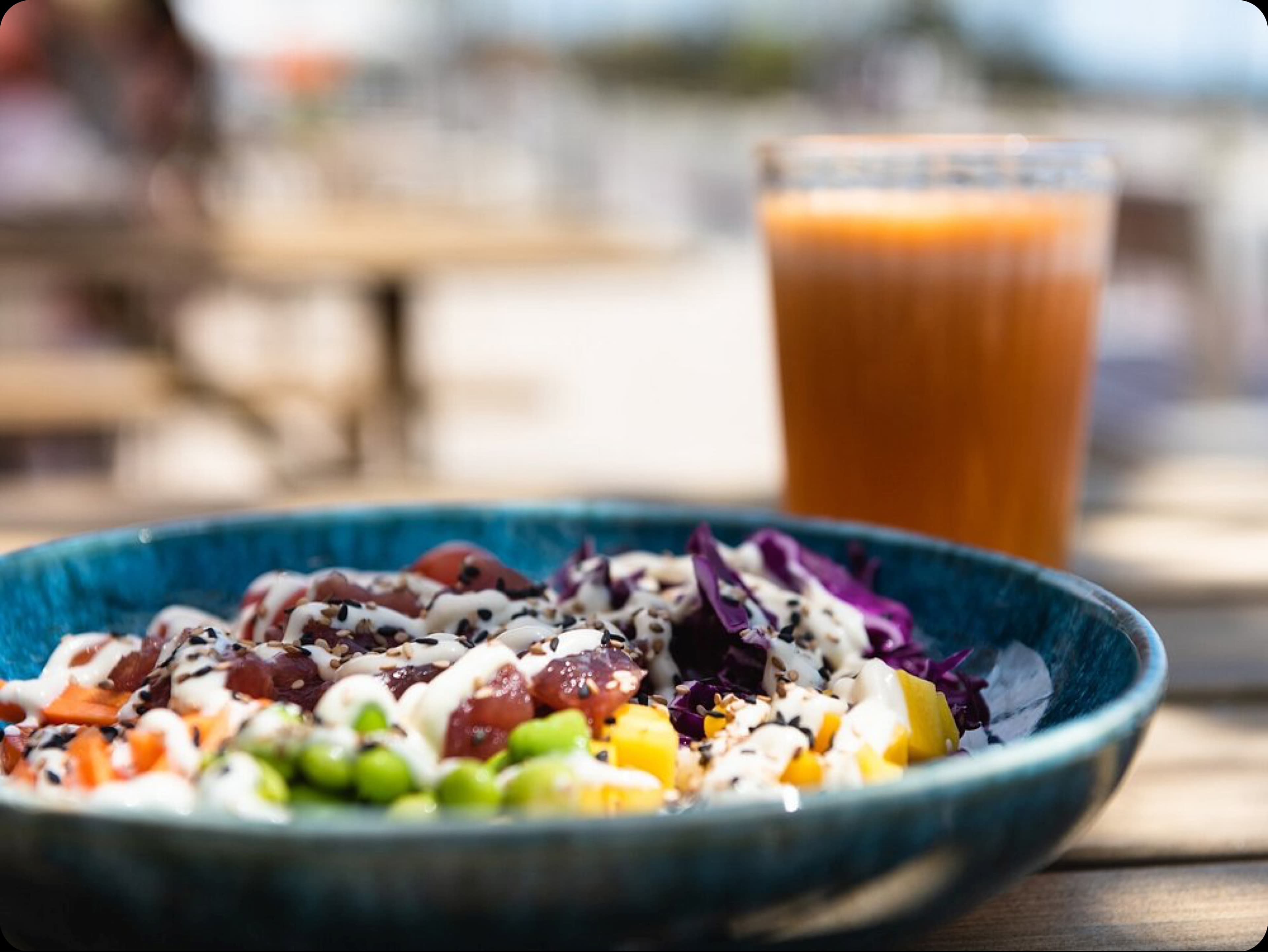 A healthy poké bowl and a glass of fresh juice served on an outdoor table with a beach view.