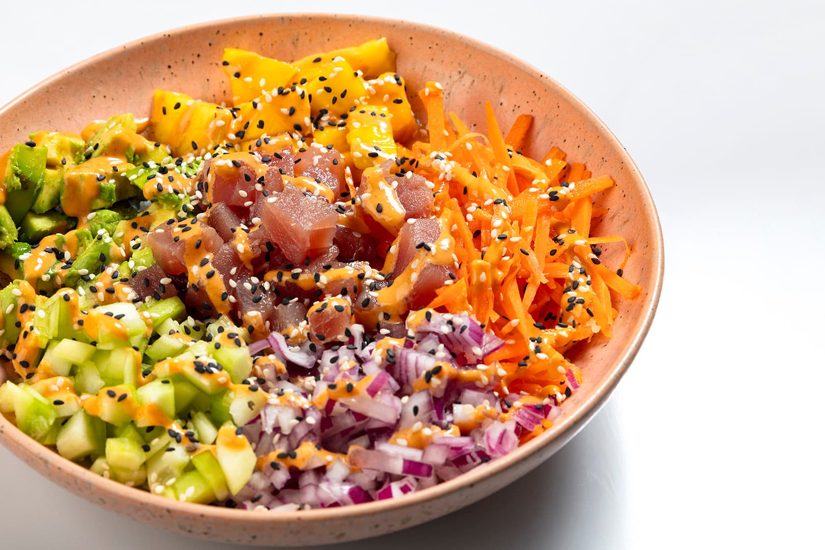 A vibrant, healthy poke bowl with carrot, mango, cucumber, onion and tuna, topped with some sauce and sesame seeds.