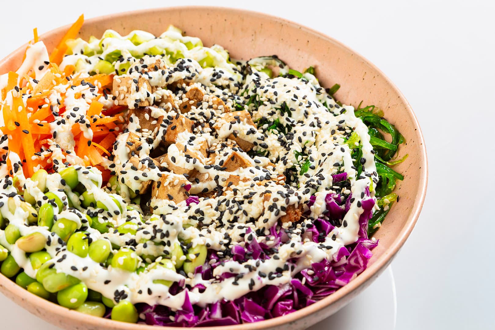A colorful poke bowl with beans, carrots, cabbage and other food topped with sauce and sesame seeds.