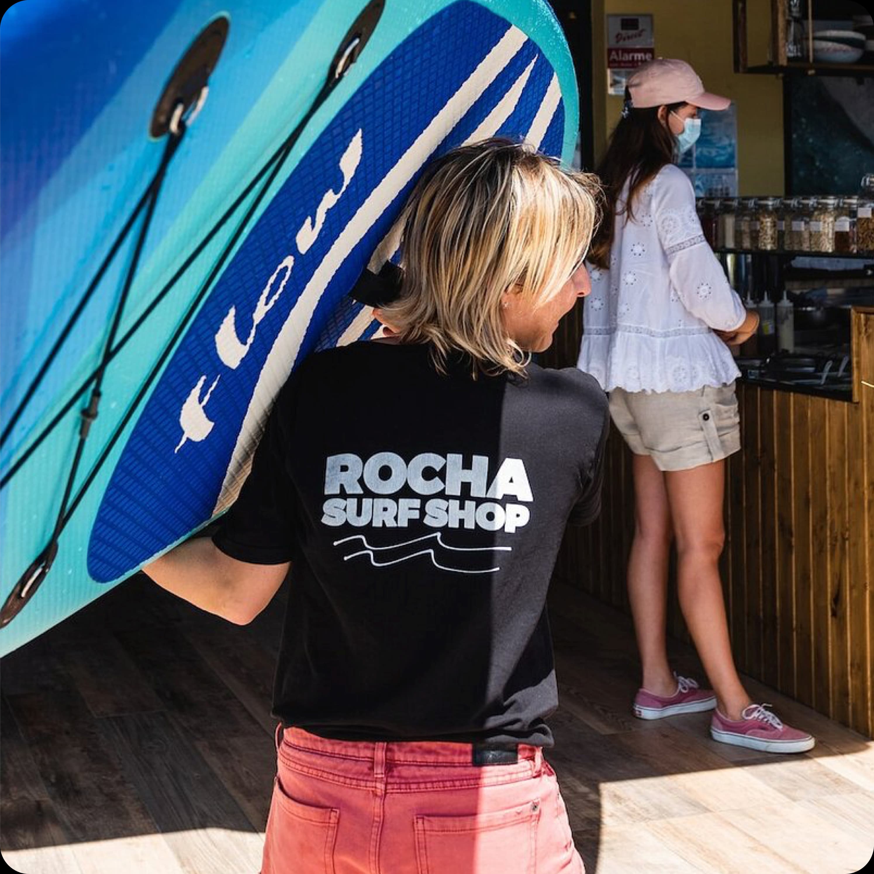 A Rocha Surf Shop team member wearing a branded t-shirt, helping a customer with a blue surfboard.