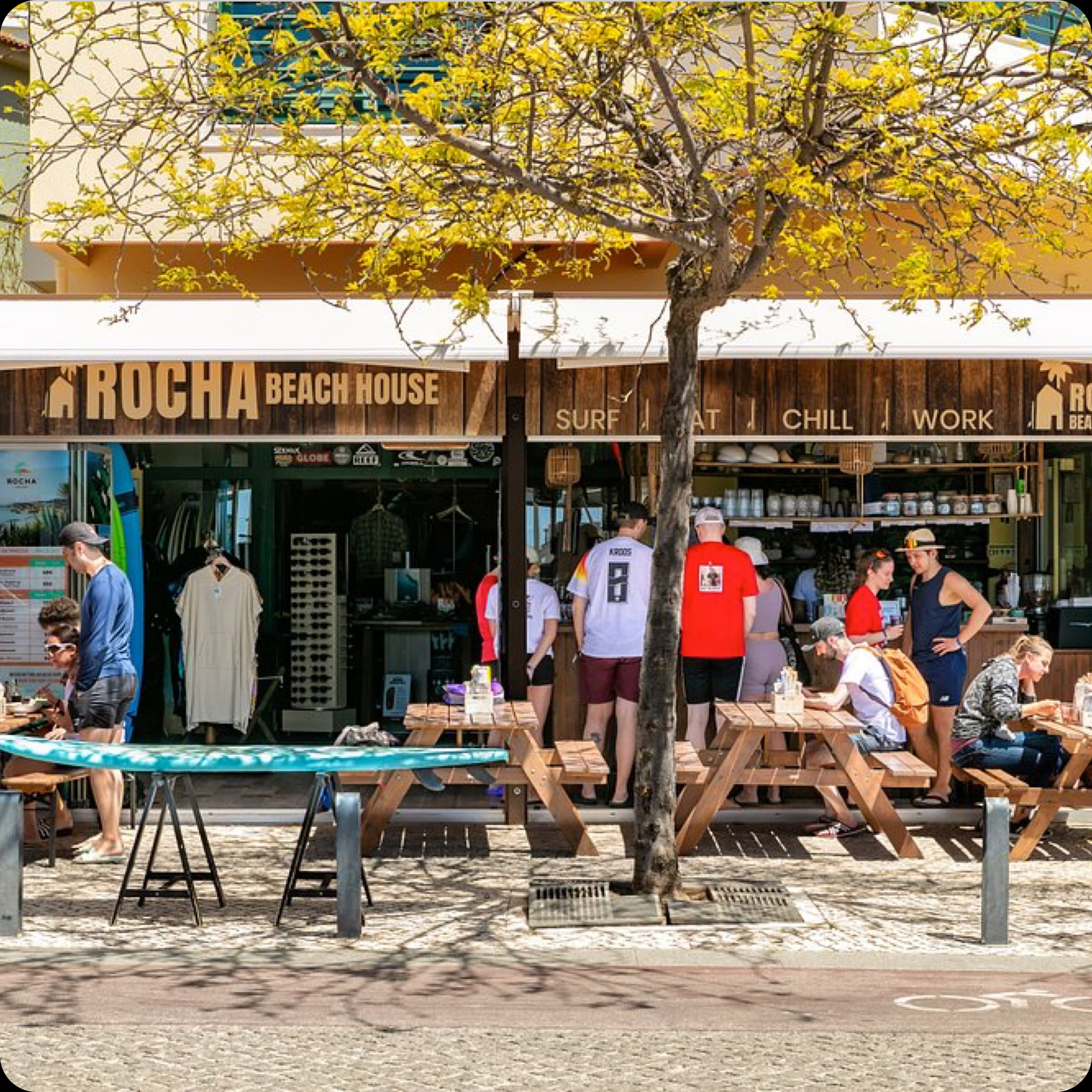 The sunny storefront of Rocha Beach House, with people enjoying the outdoor seating and surfboards on display.