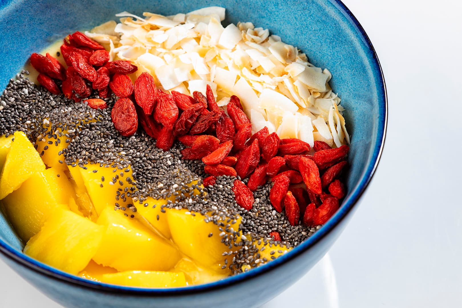 Colorful smoothie bowl with mango chunks, chia seeds, goji berries, and coconut flakes in a blue bowl.